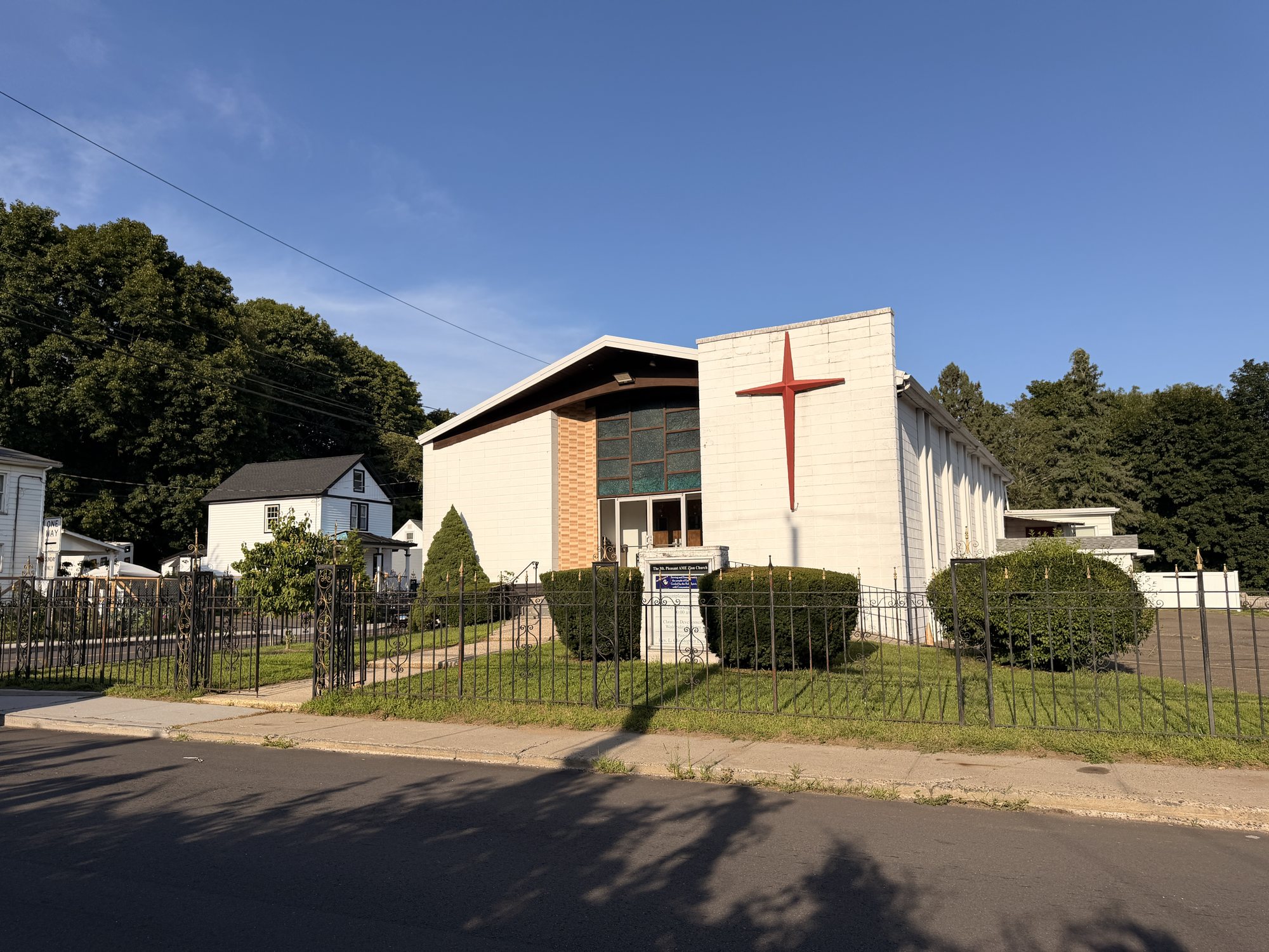 The white church building on Rowan Street with a tall red cross, stained-glass window and wrought-iron fence