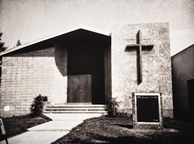 Black-and-white photo of the new church building, with a sloped roof and a tall cross on its stone front