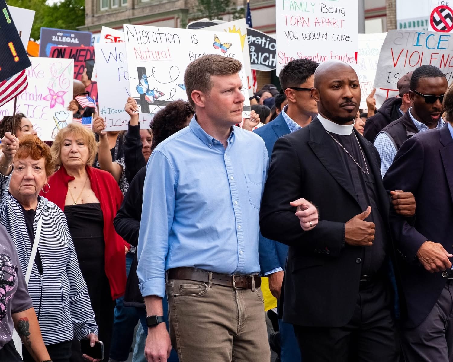 Rev. L. Christopher Lewis marching arm in arm with community members at a rally, protest signs behind them