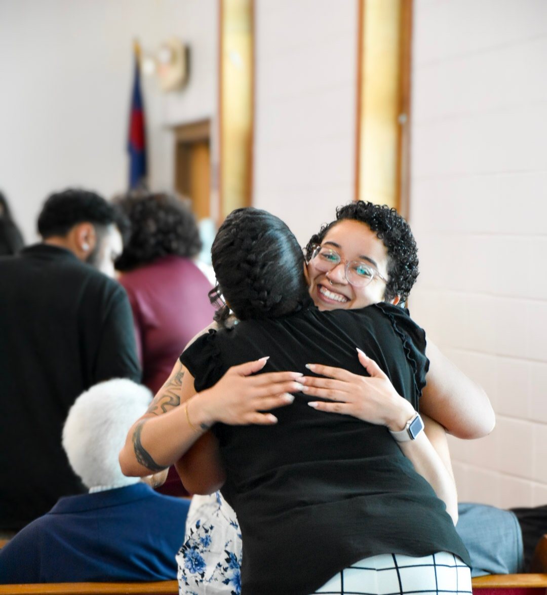 Two women embracing in the sanctuary, one smiling over the other’s shoulder